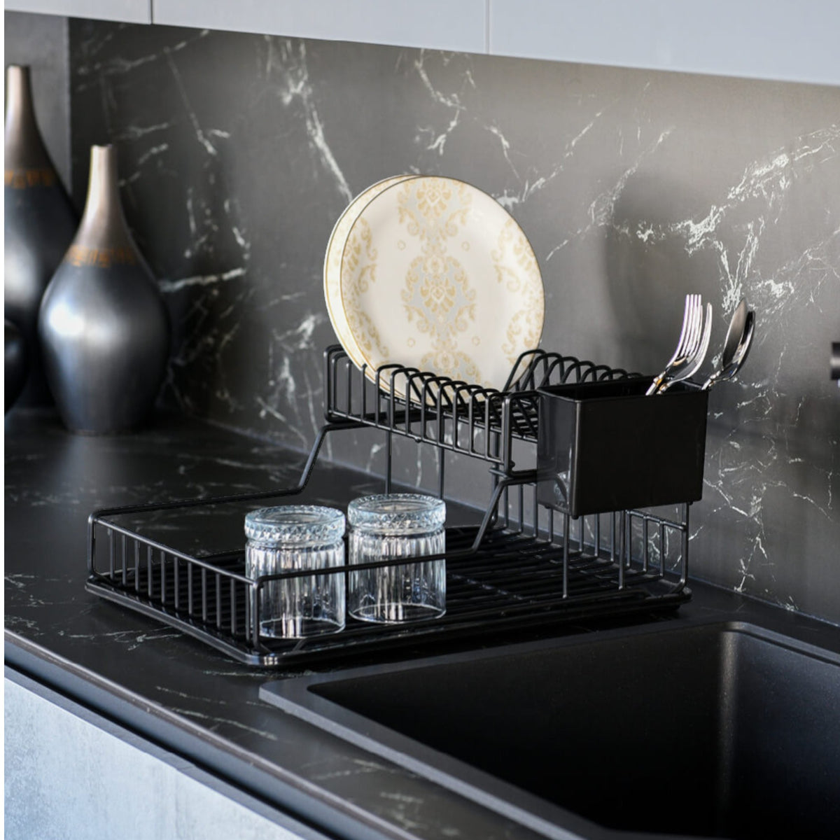 Black two-tier dish rack holding plates, glasses and cutlery beside a dark kitchen sink