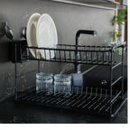 Black two-tier dish rack holding plates and glasses against a dark splashback