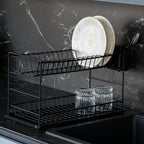 Black two-tier dish rack holding plates and glasses beside a dark kitchen sink