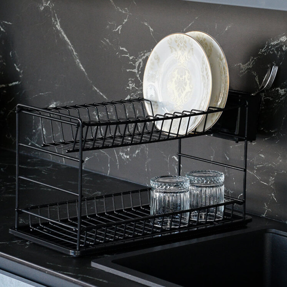 Black two-tier dish rack holding plates and glasses beside a dark kitchen sink